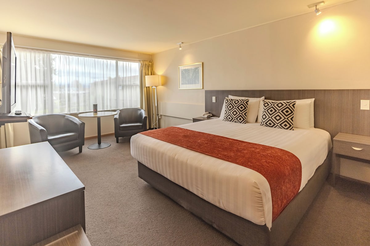 A hotel room with a large bed, a desk, a chair, a small table, and a lamp.