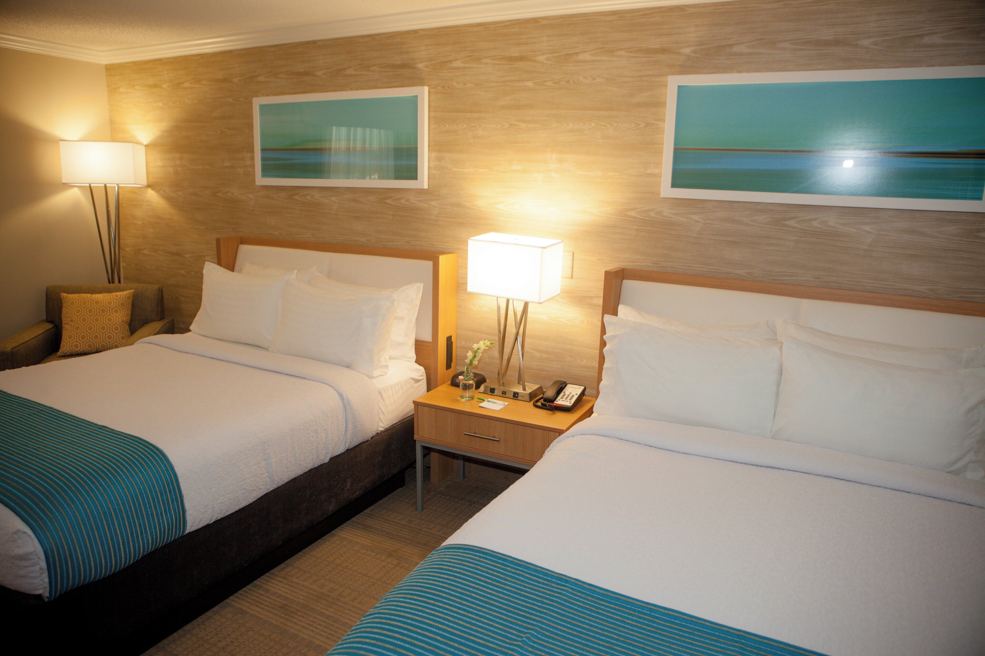 A hotel room with two beds, a nightstand with a lamp, a phone, and artwork on the wall.