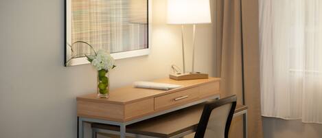 A hotel room with a desk, chair, lamp, and a painting on the wall.