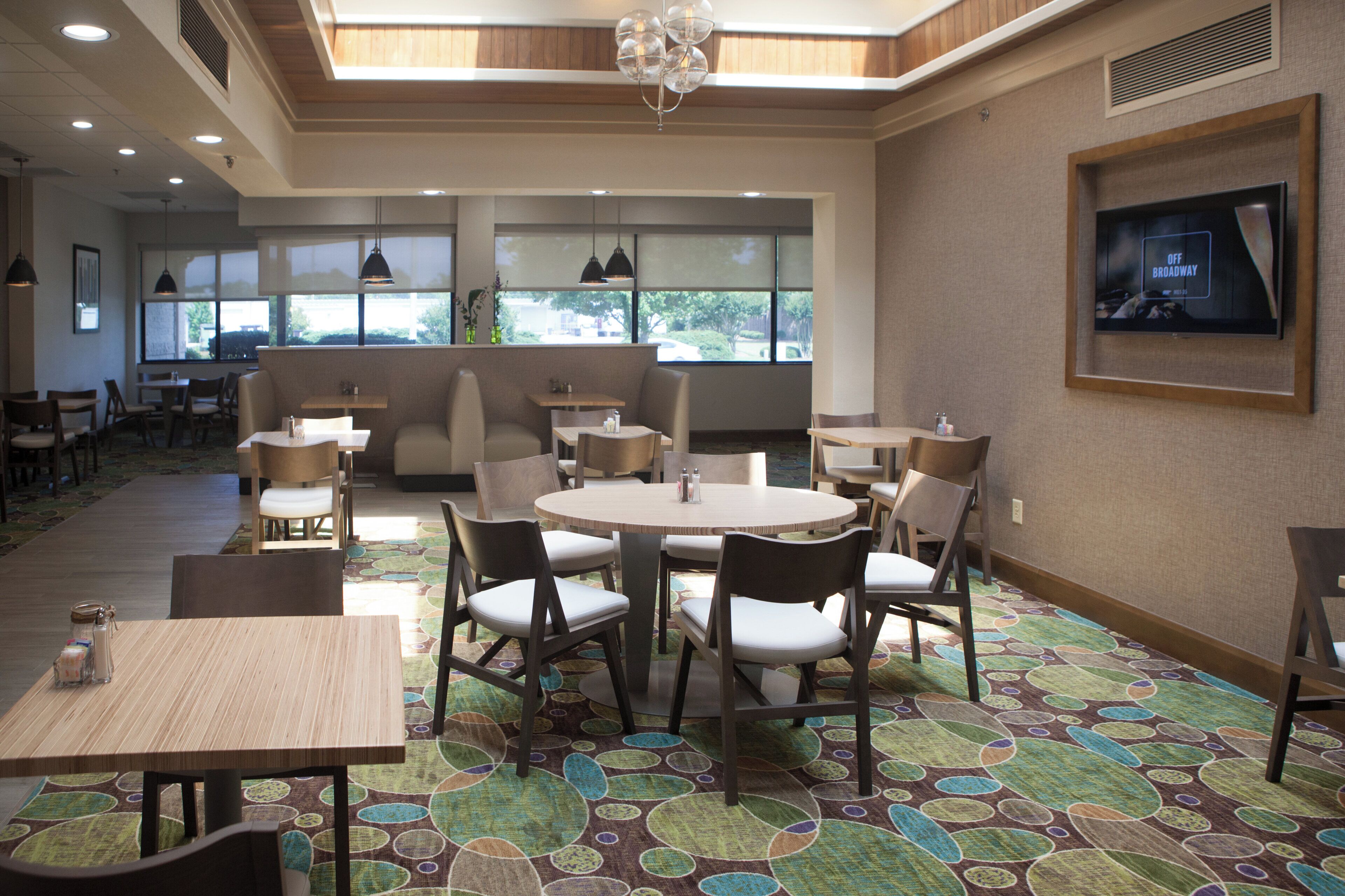A dining area with round tables and chairs, a television mounted on the wall, and a patterned carpet.