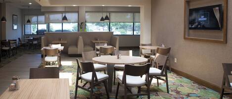 A dining area with round tables and chairs, a television mounted on the wall, and a patterned carpet.