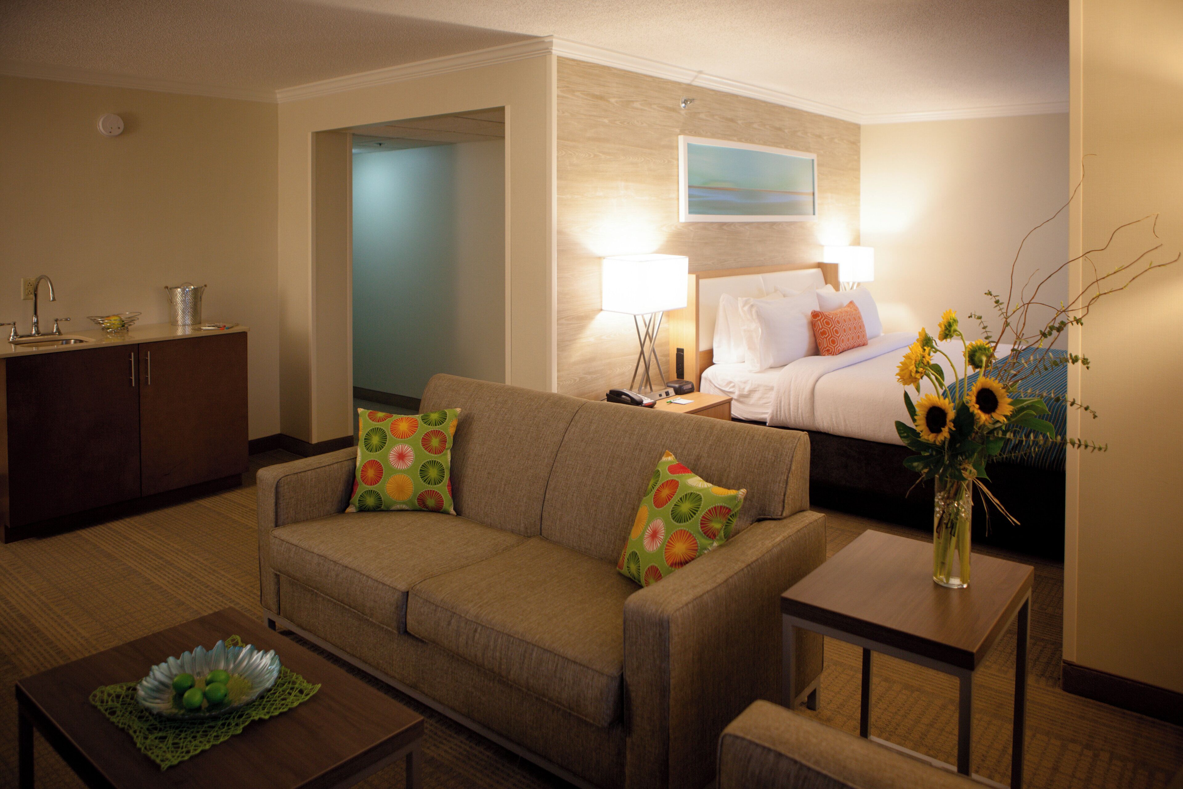 Habitación de hotel con sofá, una mesita, una mesita de noche, lámpara, cama, un cuadro en la pared y un jarrón con flores.
