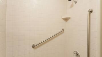 Un baño con paredes de azulejos blancos, un cabezal de ducha manual, una barra de apoyo y un estante en la esquina.