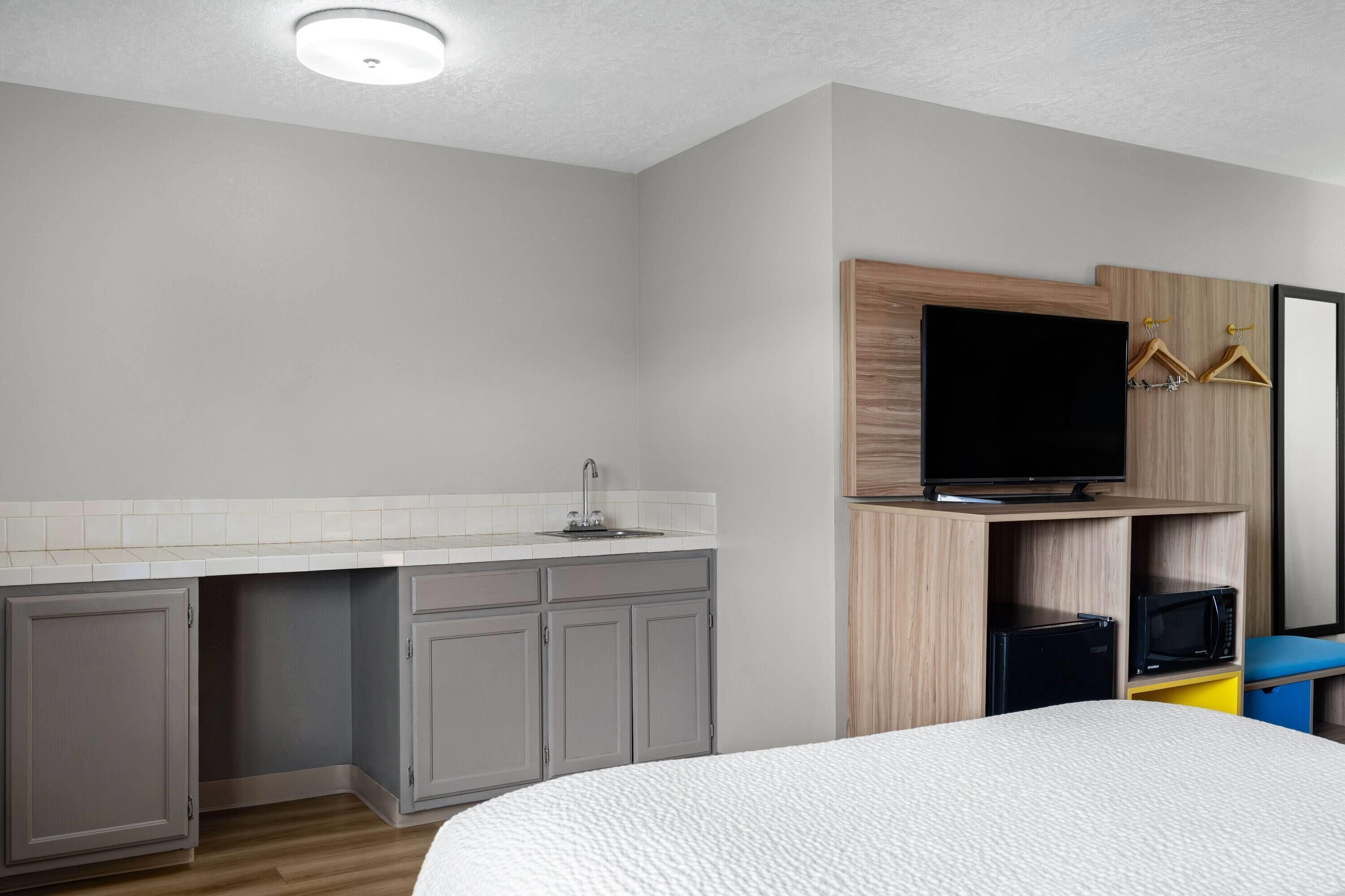 A compact hotel room with a kitchenette, a bed, a flat-screen TV, and a small refrigerator.