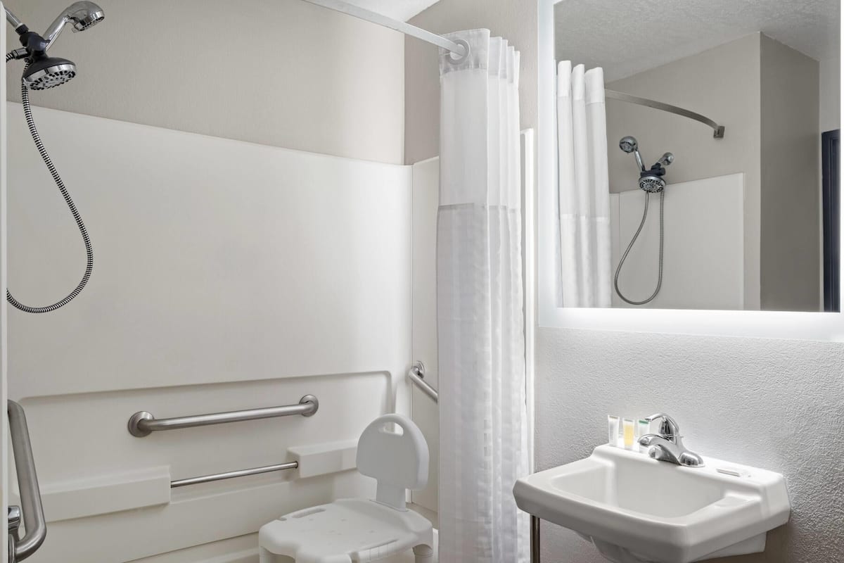 A bathroom with a shower, a toilet with a safety bar, a sink, and a mirror.