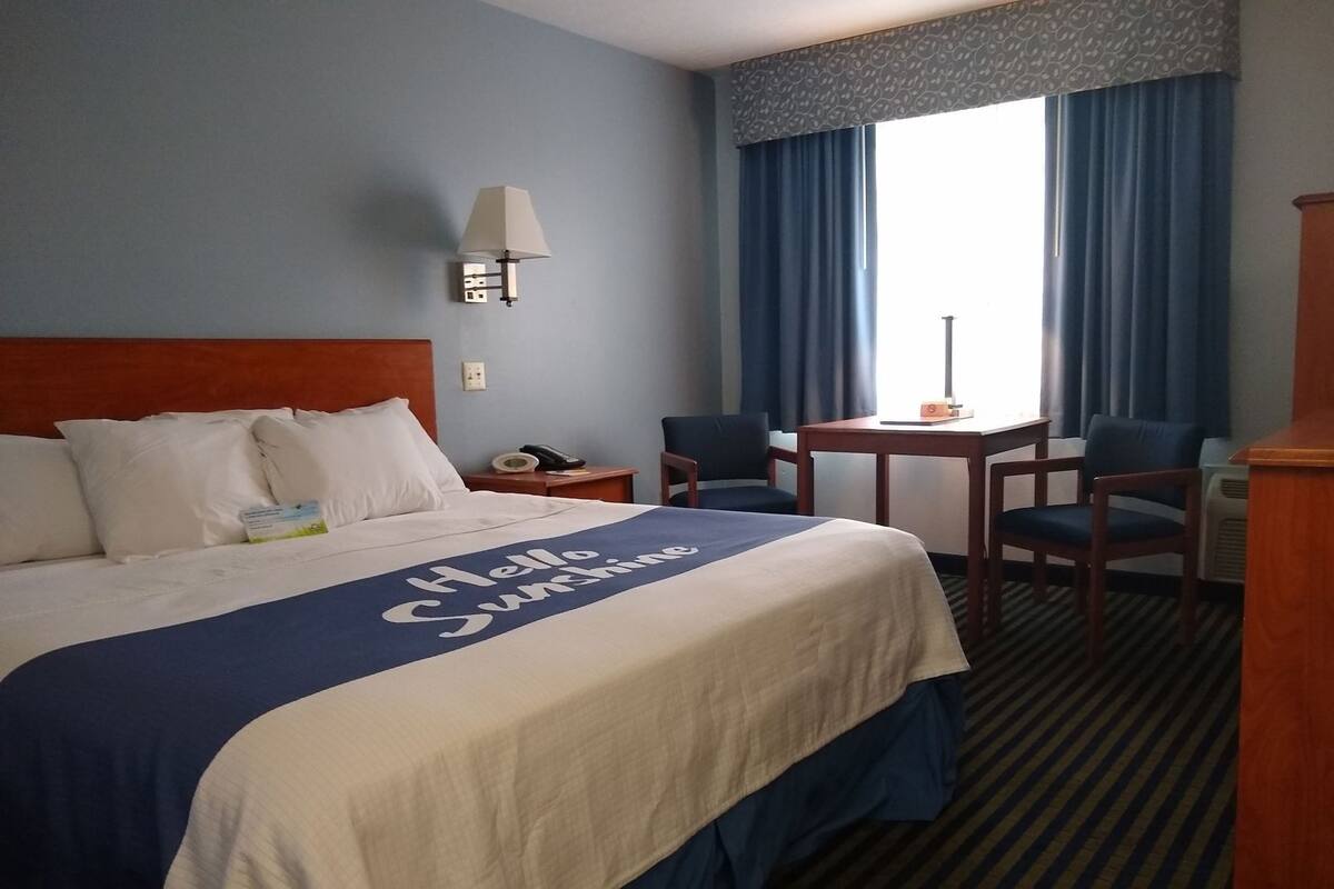 A hotel room with a bed, a desk, two chairs, a lamp, and a window with curtains.