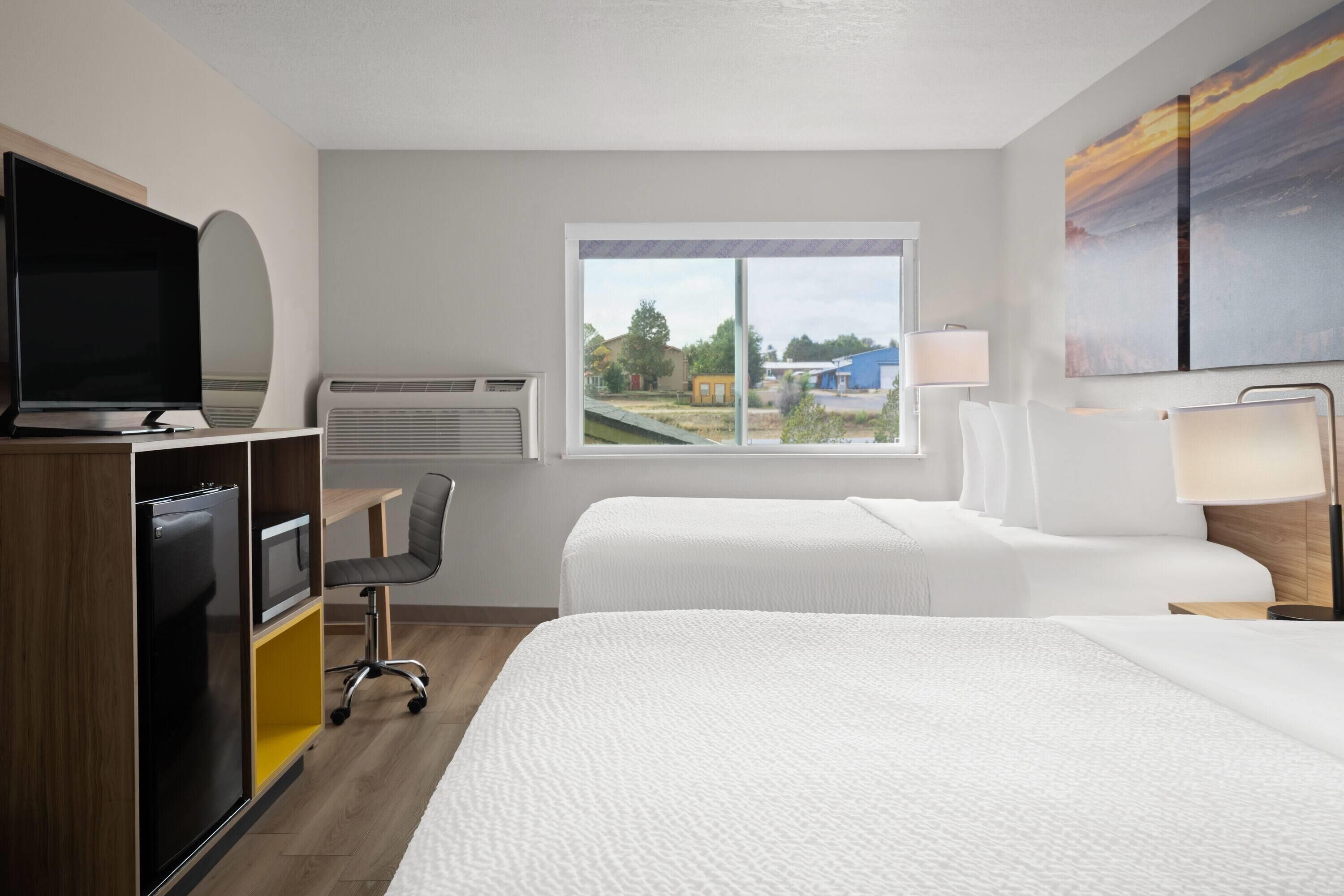 A hotel room with two beds, a desk, a chair, a TV, and a window with a view of buildings.