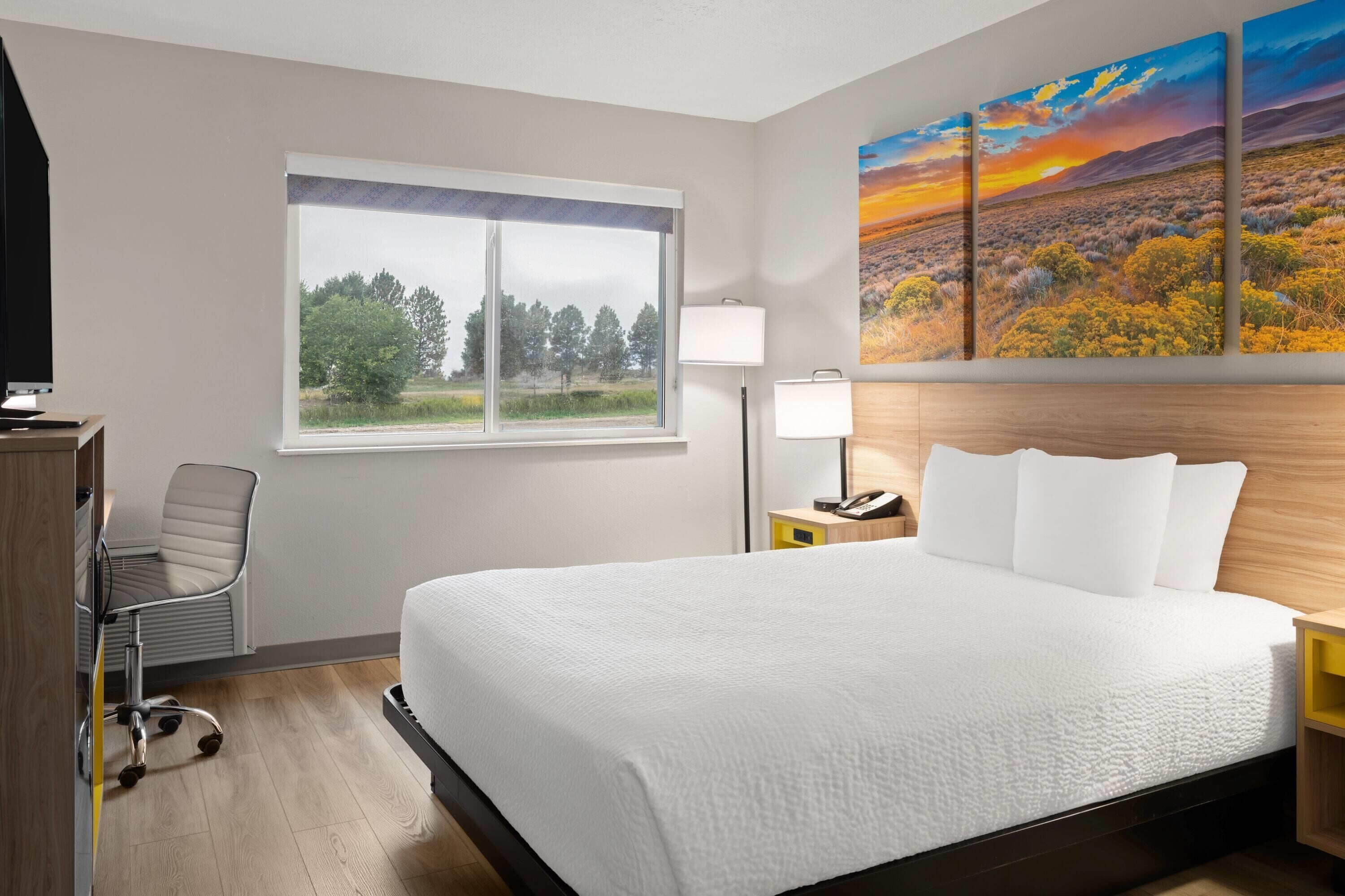 A hotel room with a large bed, a desk, a chair, a window with a view of trees, and a painting of a sunset landscape.