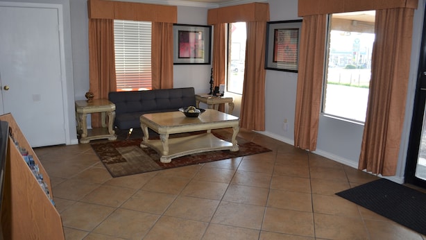 A living room with a sofa, coffee table, and two side tables. There are curtains on the windows, and a door is visible on the left. - Hill Country Inn & Suites (Copperas Cove)