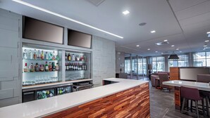 A modern bar area with a wooden counter, glass shelves with bottles, and seating in the background.