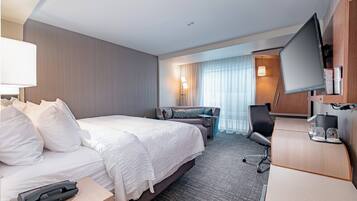 A hotel room with a bed, bedside table, lamp, TV, desk, chair, and a sofa.