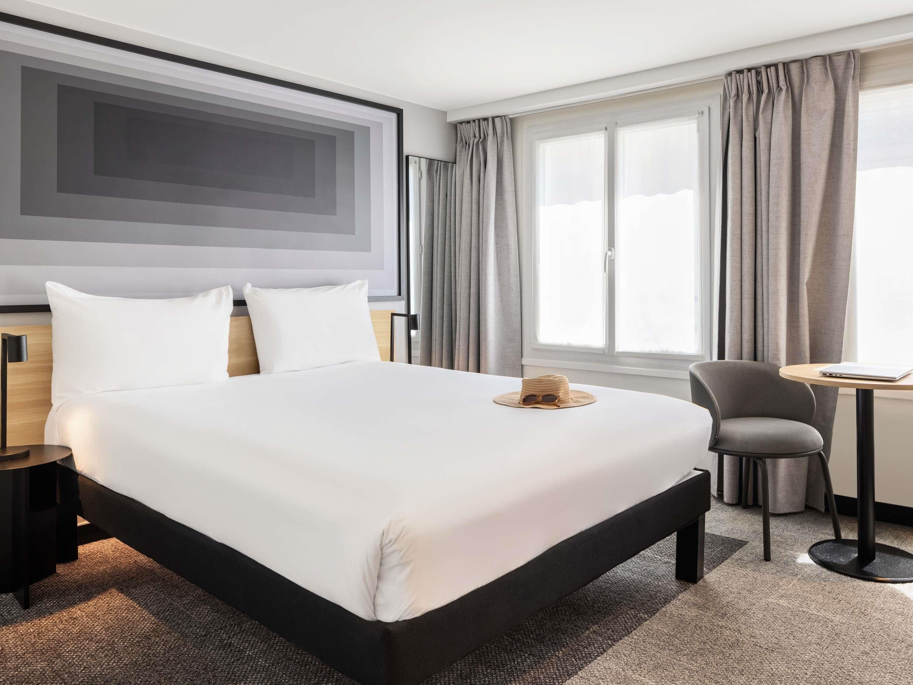 A modern hotel room with a large bed, a desk, a chair, and a view of the outdoors.