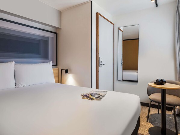 Une chambre d’hôtel avec un lit, une petite table et une chaise, un miroir et une fenêtre.