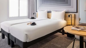 A hotel room with two beds, a bedside table, a lamp, and a window with curtains. - Ibis Paris Grands Boulevards Opera 9th (Paris)
