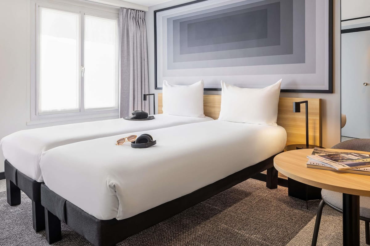 A hotel room with two beds, a bedside table, a lamp, and a window with curtains.