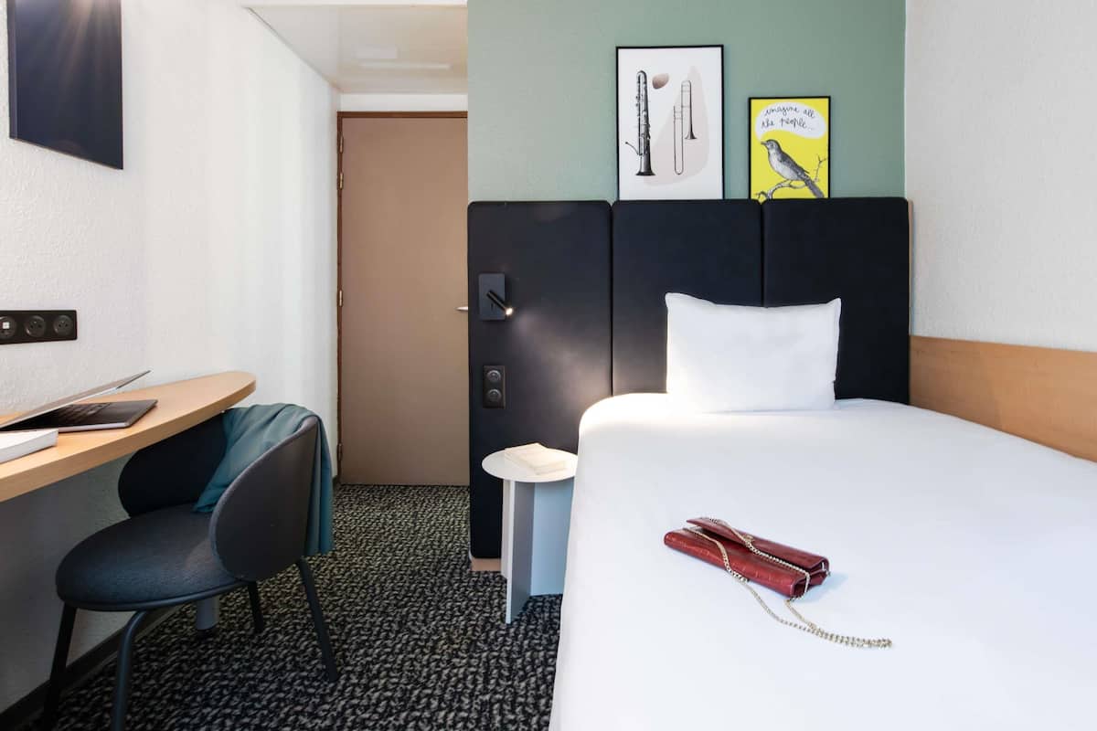 A hotel room with a bed, a desk with a laptop, a chair, a small side table, and a door.