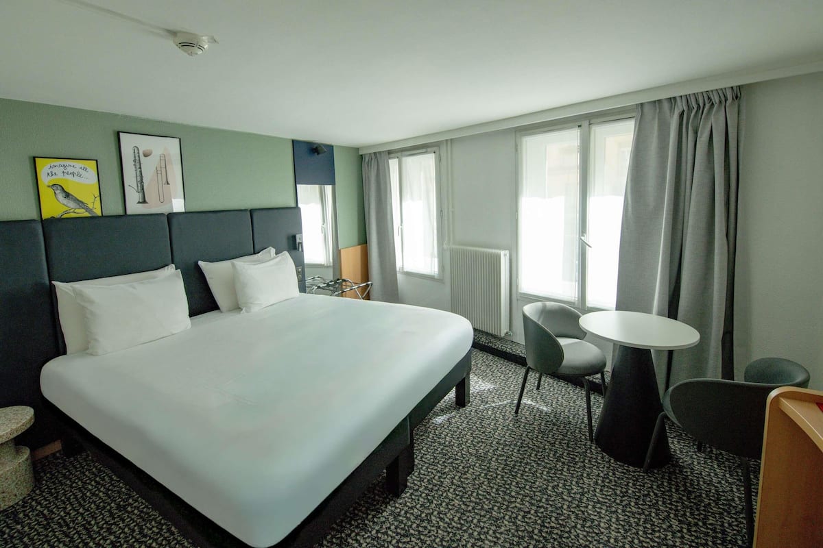 A hotel room with a large bed, a small table, two chairs, a radiator, and a window with curtains.
