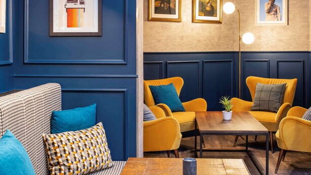 A modern lounge area with blue and yellow furniture, framed artwork on the walls, and pendant lighting. - Ibis Paris Grands Boulevards Opera 9th (Paris)