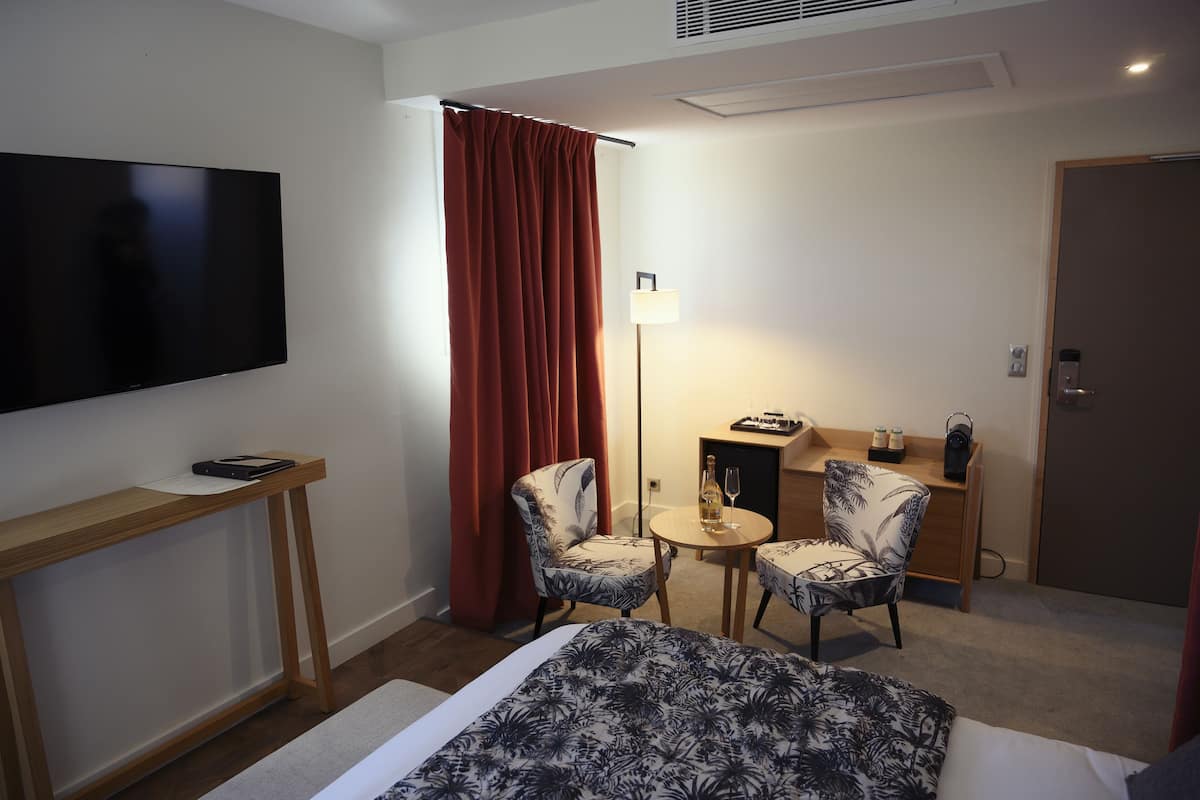 Une chambre d’hôtel avec un lit, deux chaises, une petite table, un téléviseur, une lampe, un bureau et une porte.