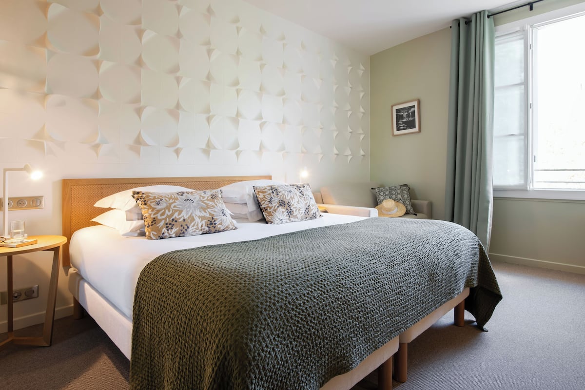 Un lit bien fait, recouvert d’une couverture verte, une table de chevet avec une lampe et un mur orné d’un motif géométrique.