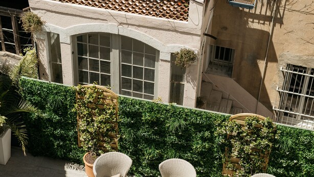 Un patio exterior con mobiliario de mimbre, un seto verde y un edificio con tejado de azulejos y ventanas.