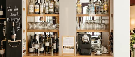 Un espace bar avec des étagères remplies de bouteilles d’alcool et de verres à vin, une machine à café et un tableau affichant le menu.