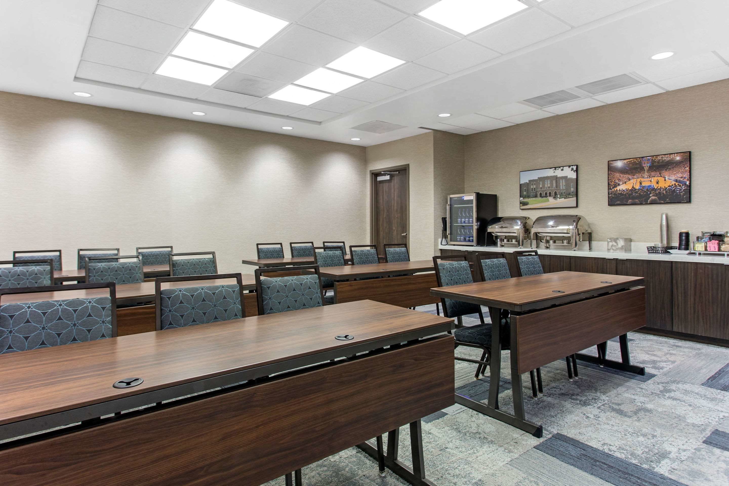 A conference room with multiple tables and chairs, a kitchenette, and two framed pictures on the wall.