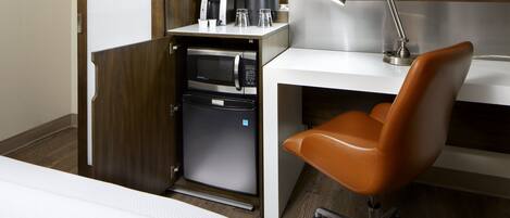 A compact hotel room with a bed, a desk with a computer, a microwave, a coffee maker, and a lamp.