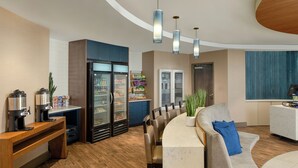 A modern hotel lobby with a curved reception desk, seating area, coffee machines, refrigerator, and a large window with a view. - The Lodge at Duke Medical Center (Durham)
