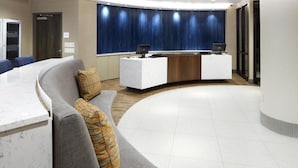 A modern reception area with a curved reception desk, a grey sofa with a patterned cushion, and a large window with a blue curtain backdrop. - The Lodge at Duke Medical Center (Durham)