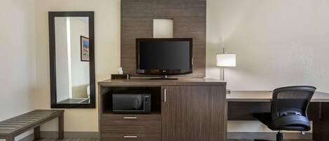 A hotel room with a wooden desk, a television, a microwave, a chair, and a bench.