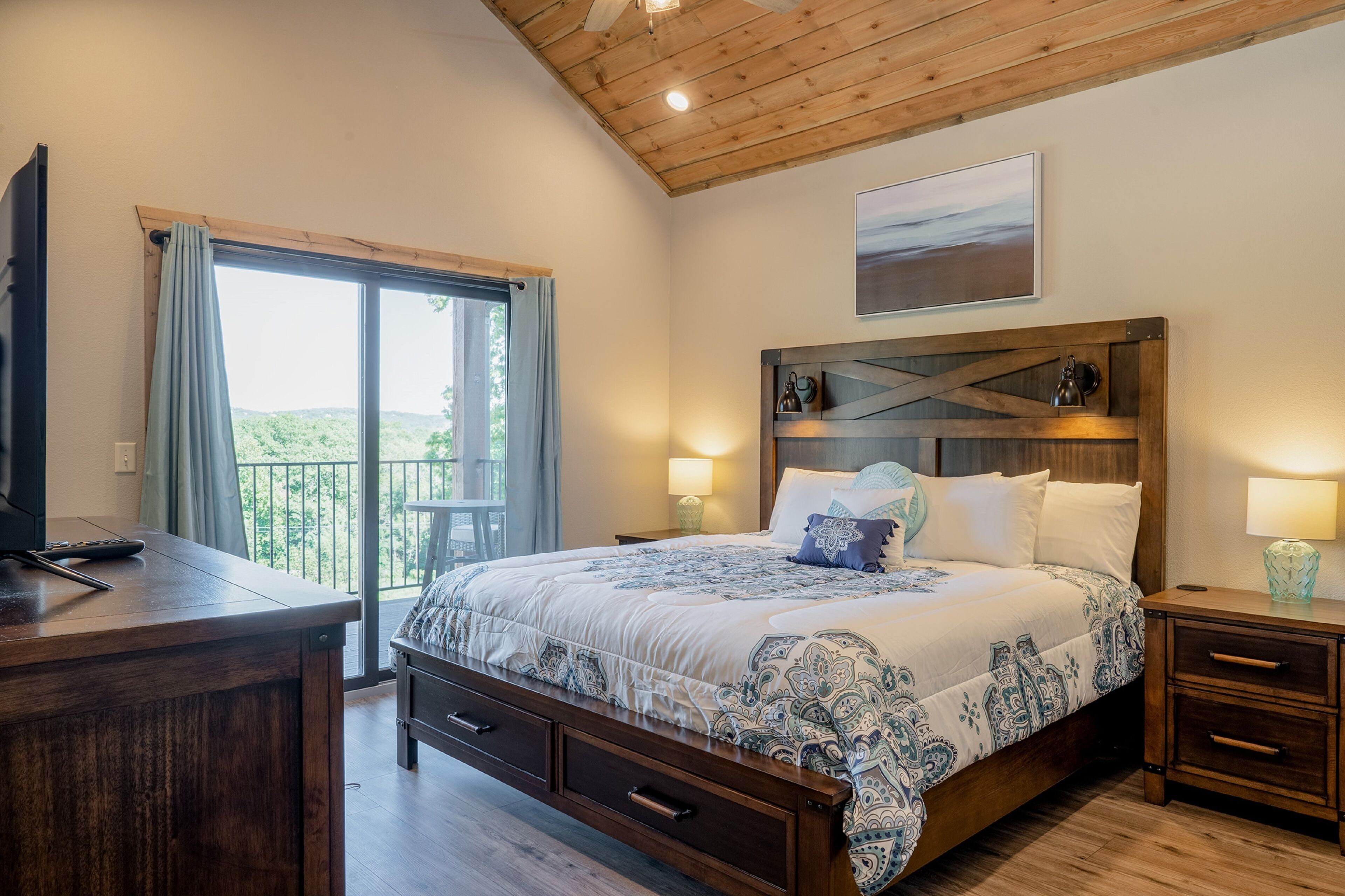 Un dormitorio con techo de madera, una cama grande con ropa de cama estampada, lámparas de noche y vistas a través de una ventana.