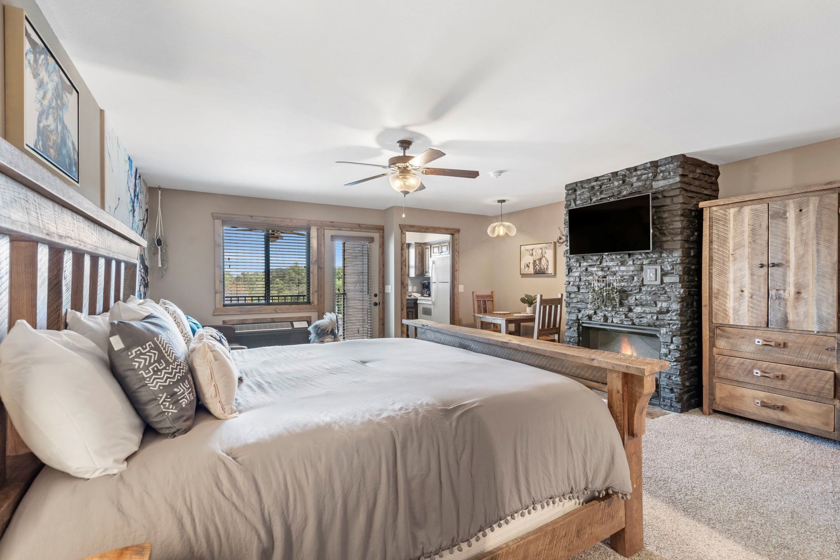 A bedroom with a large bed, a wooden bench, a stone fireplace, and a TV mounted on the wall.