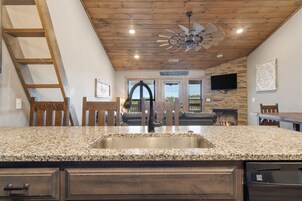 A kitchen with granite countertops, a stainless steel sink, wooden cabinets, a ceiling fan, and a stone wall.