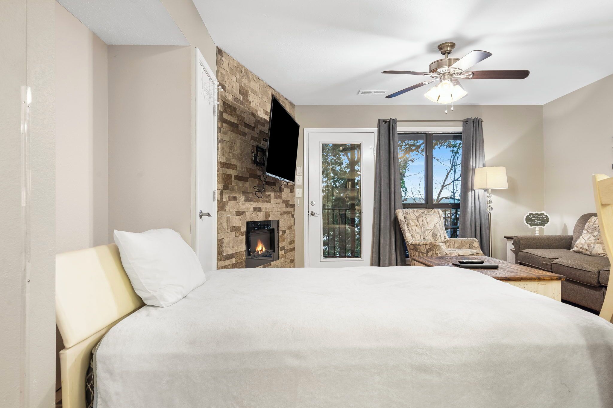 A bedroom with a bed, a fireplace, a ceiling fan, a TV, and a view of the outdoors.