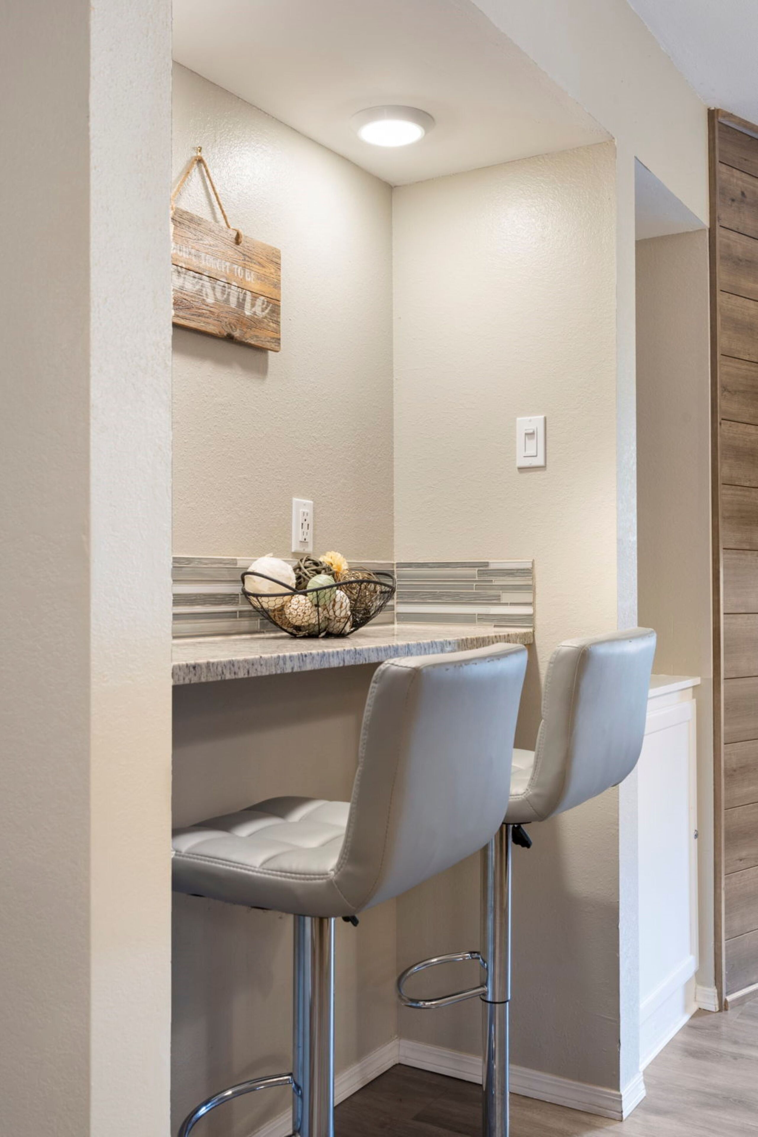 A corner kitchenette with two bar stools, a small countertop with a basket of fruits, and a hanging wooden sign.