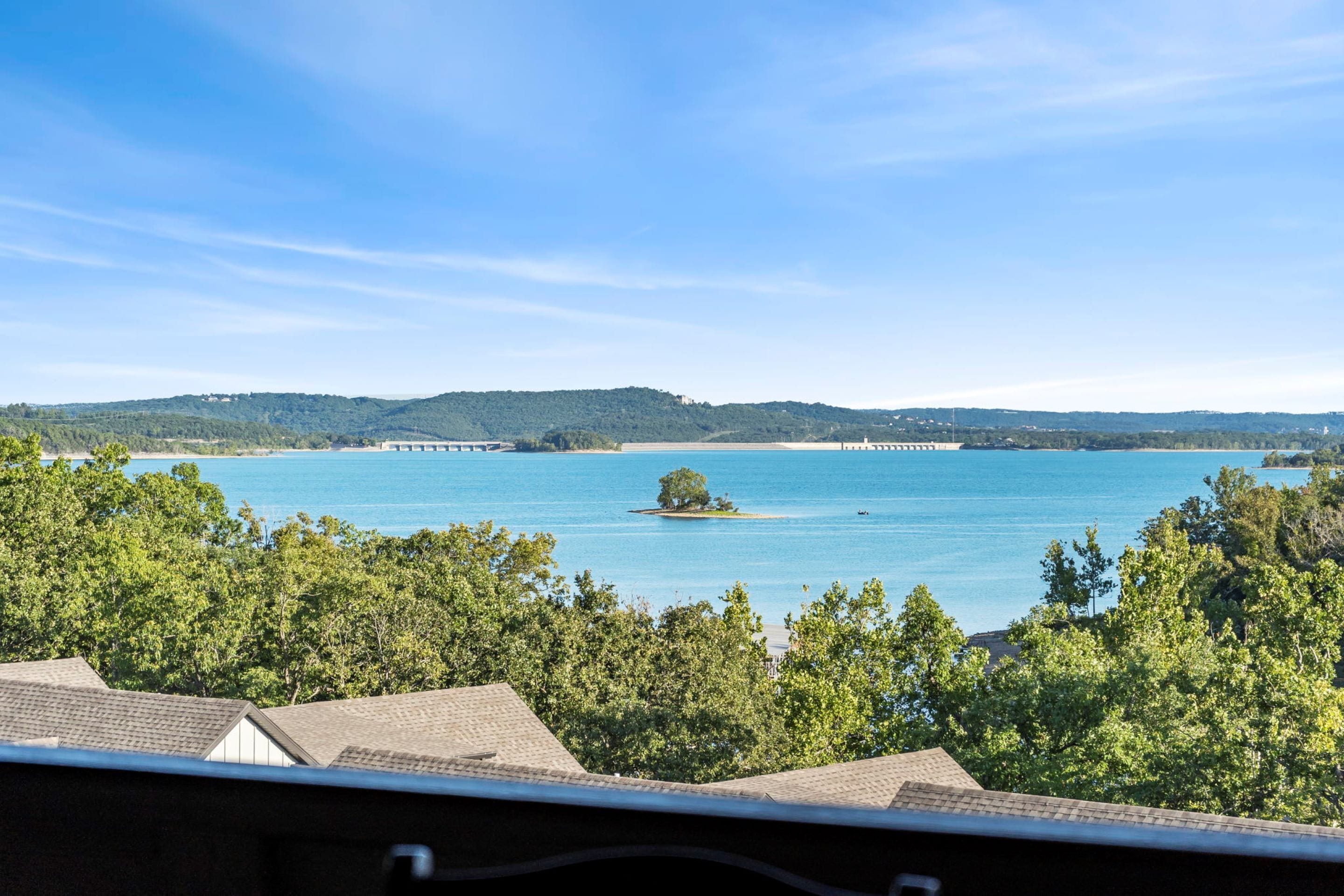 Blick auf einen See mit einer kleinen Insel, umgeben von Bäumen und Hügeln im Hintergrund.
