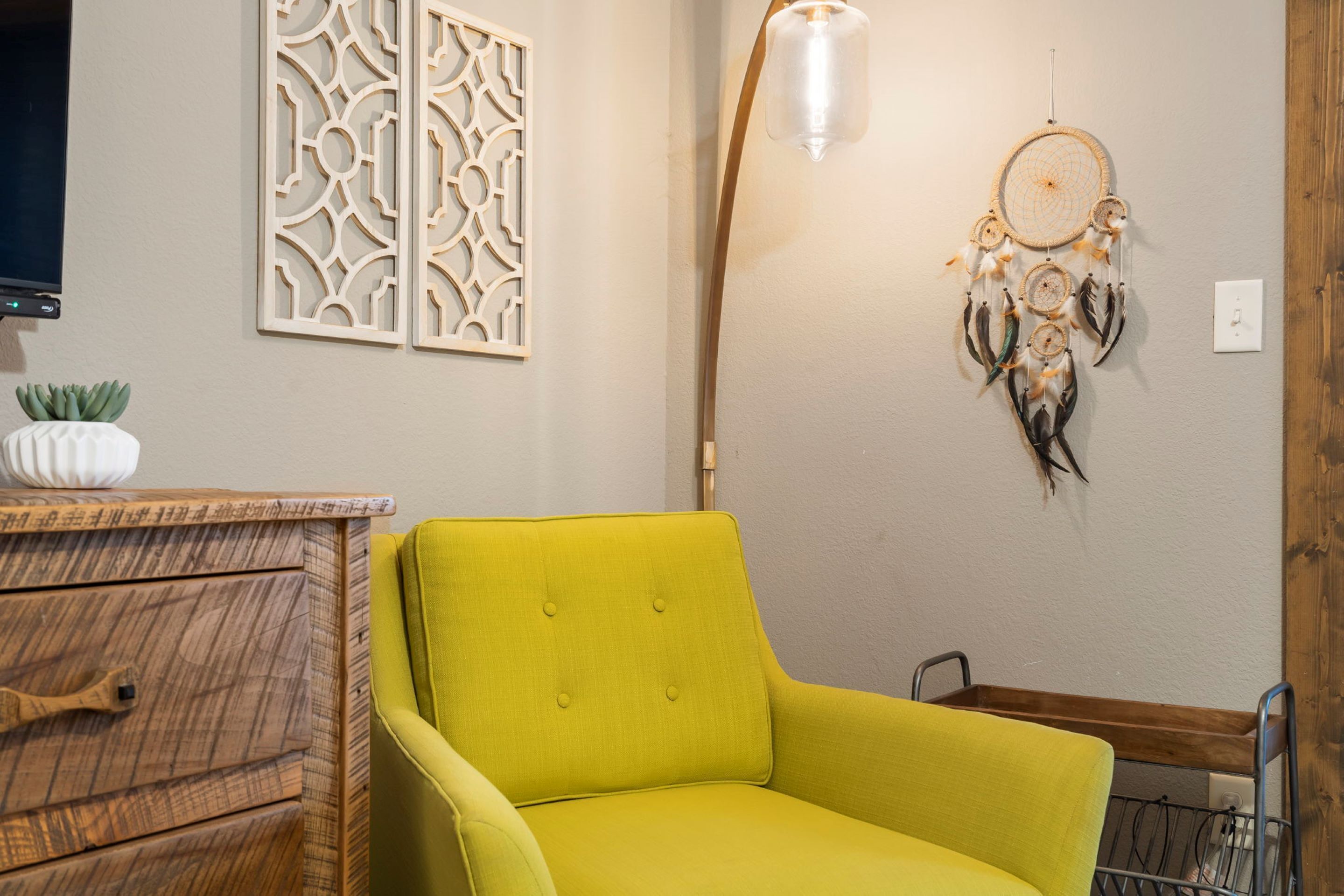Un sillón amarillo brillante en una habitación con un panel decorativo, un atrapasueños y un aparador de madera.