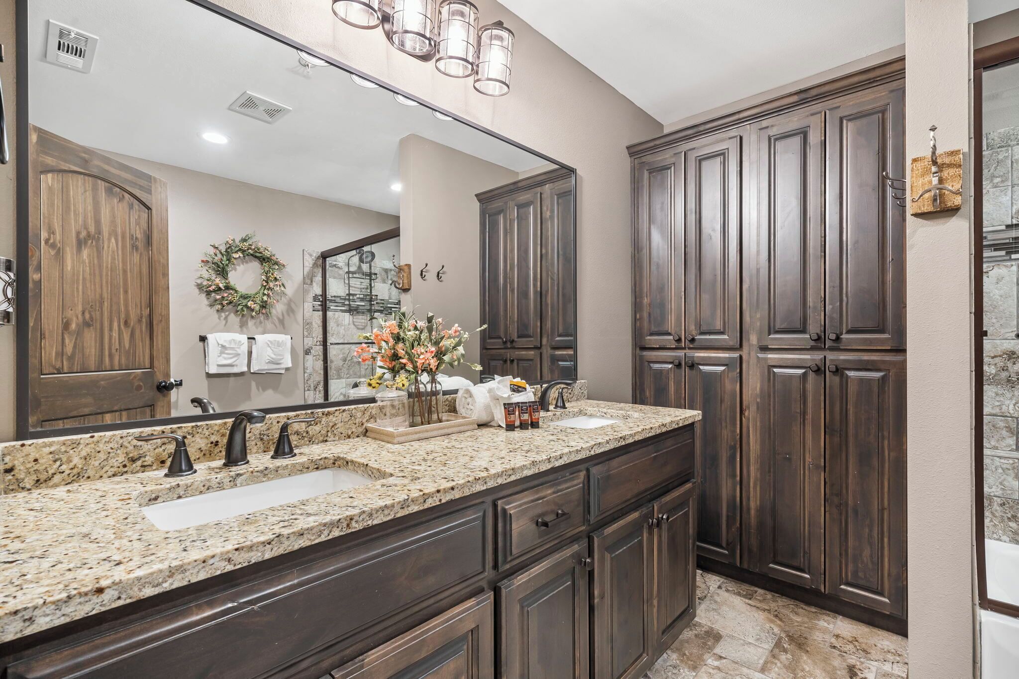 A bathroom with a granite countertop, dark wooden cabinets, a large mirror, and a shower area.