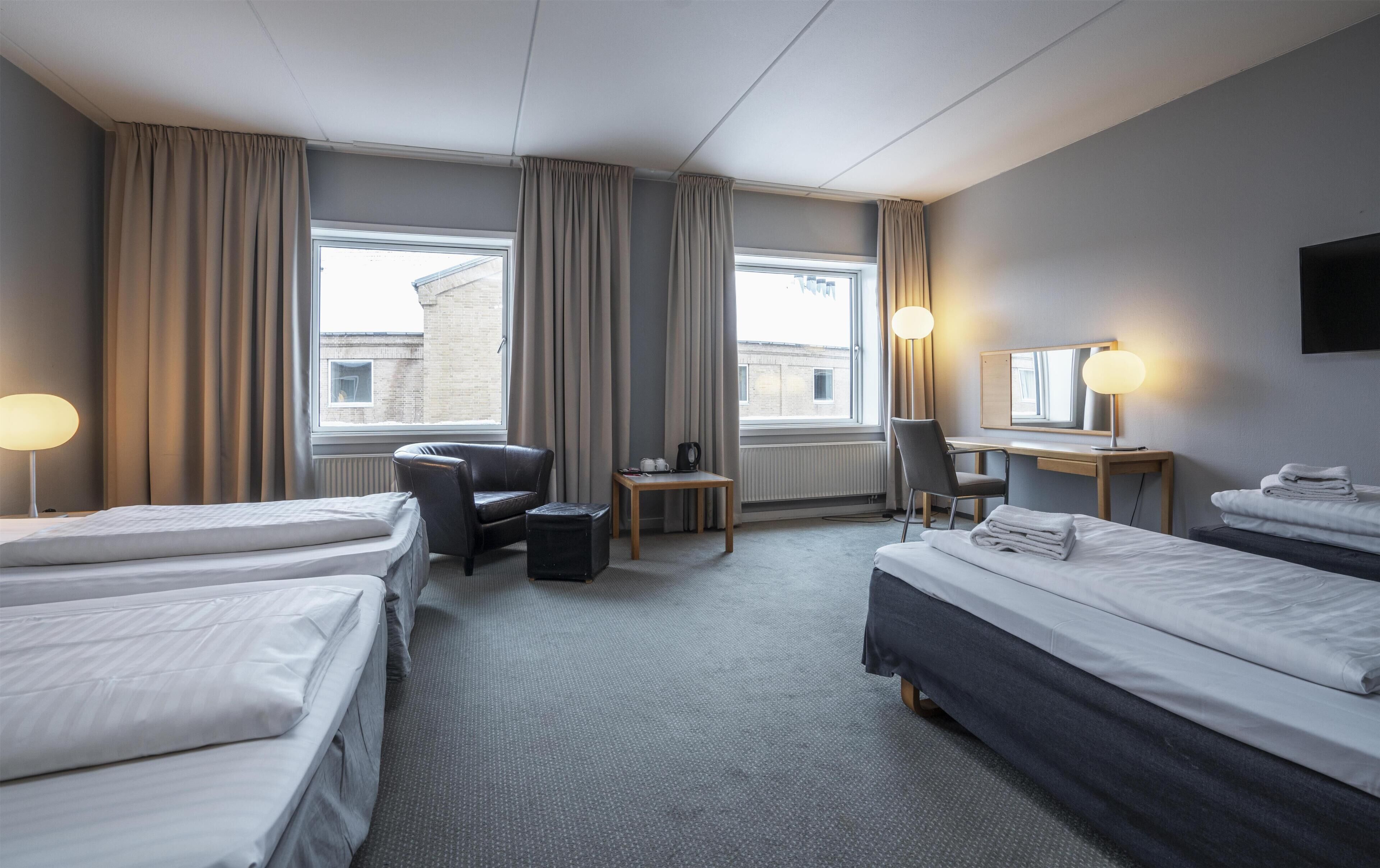 A hotel room with two beds, a desk, a chair, a small table, and a radiator.
