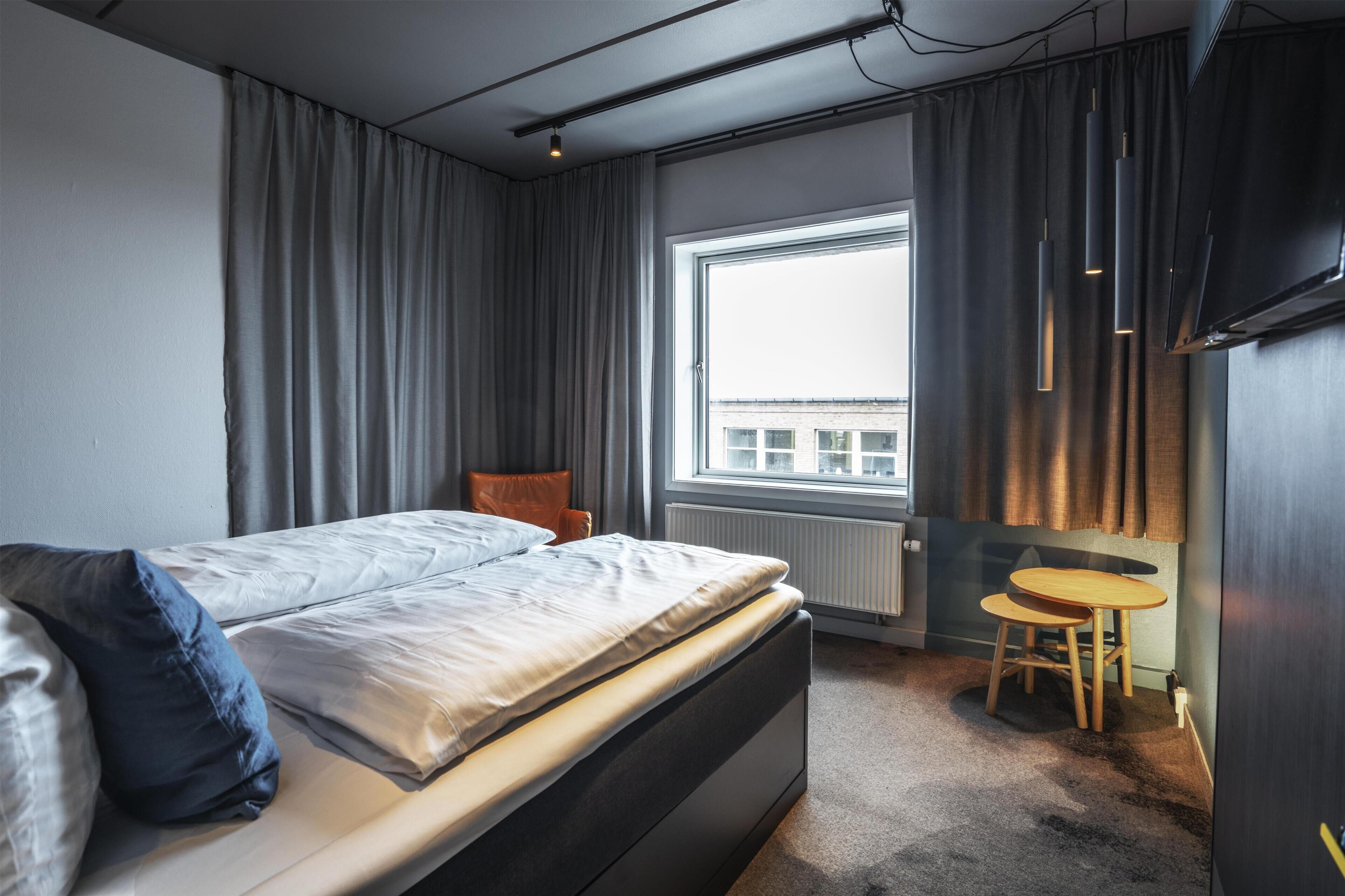 Une chambre d’hôtel moderne équipée d’un lit, d’une petite table, d’une chaise, d’une fenêtre avec des rideaux et d’un téléviseur.