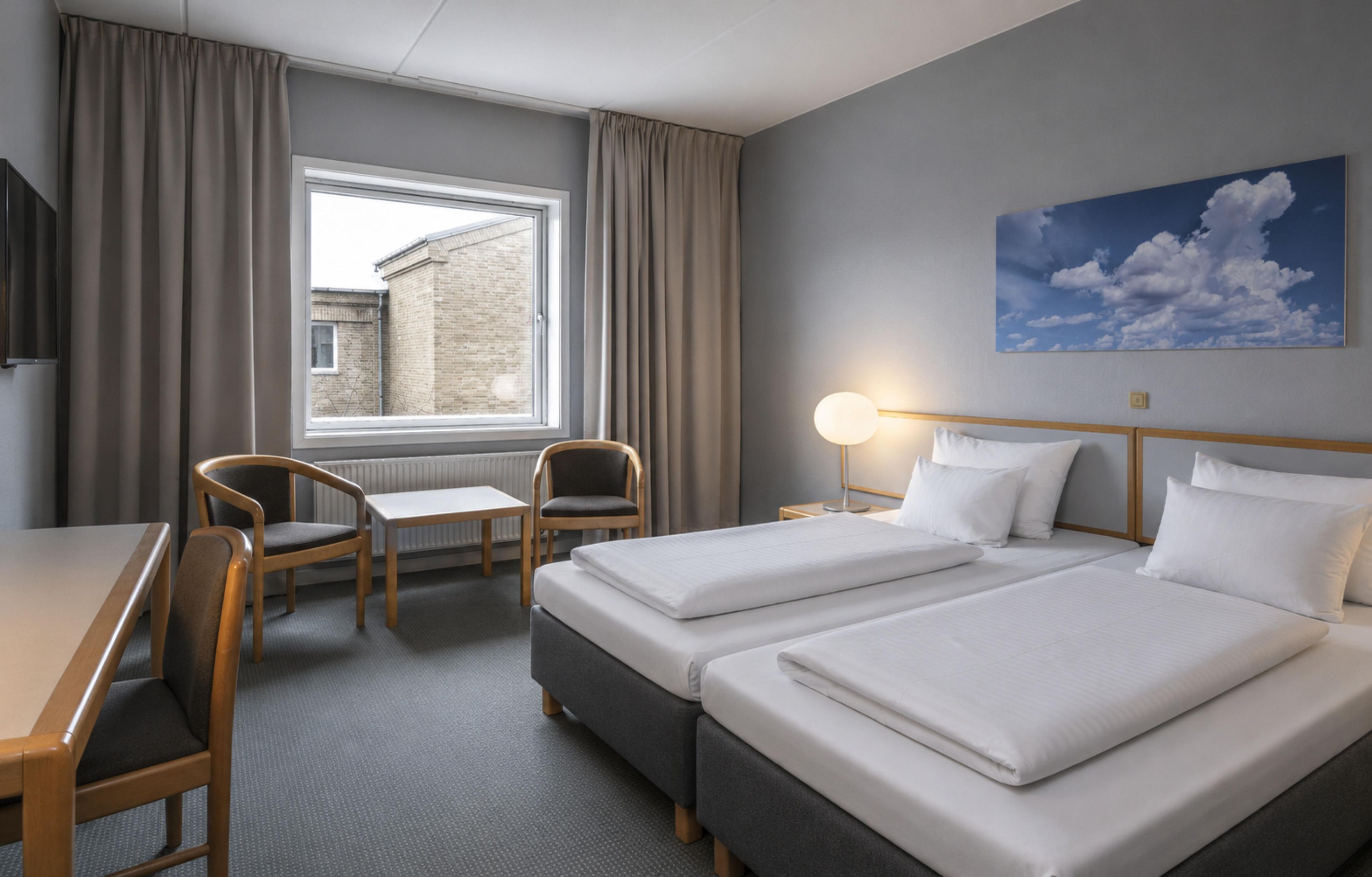 Habitación de hotel con dos camas, un escritorio, dos sillas, una ventana con cortinas, una lámpara y un cuadro de nubes.