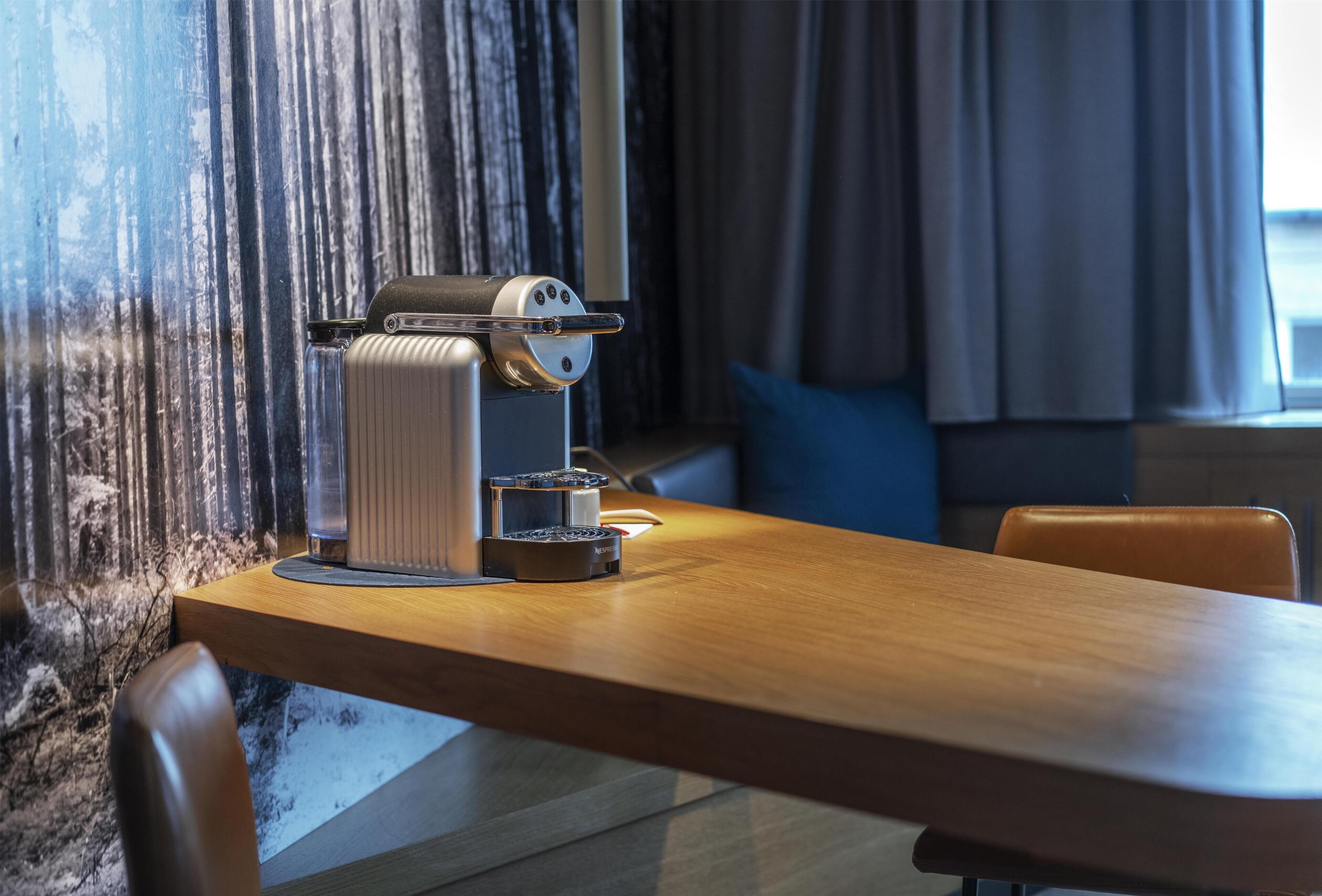 Een koffieapparaat op een houten tafel in een kamer met een gestructureerde muur en een raam met gordijnen.