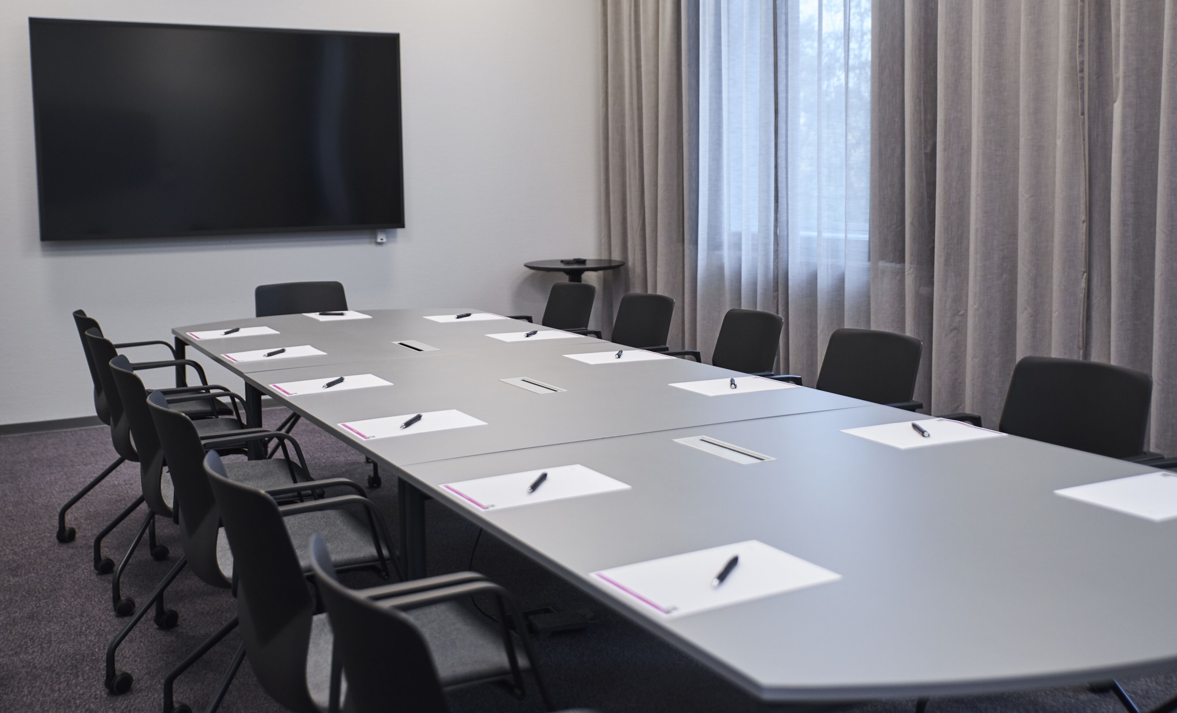 Ein Konferenzraum mit langem Tisch, Stühlen, einer Projektionsleinwand und einem Fenster mit Vorhängen.