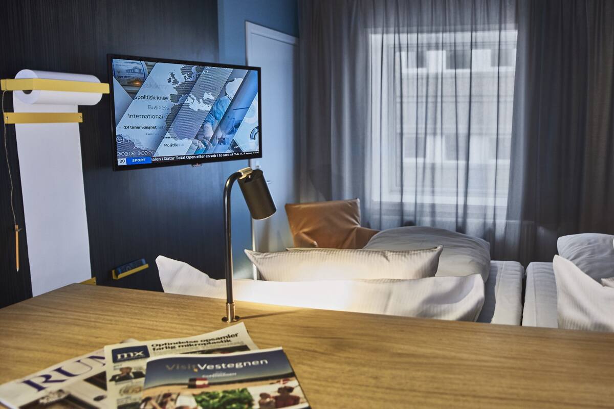 A hotel room with a bed, a desk with a lamp, a TV on the wall, and a window with curtains.