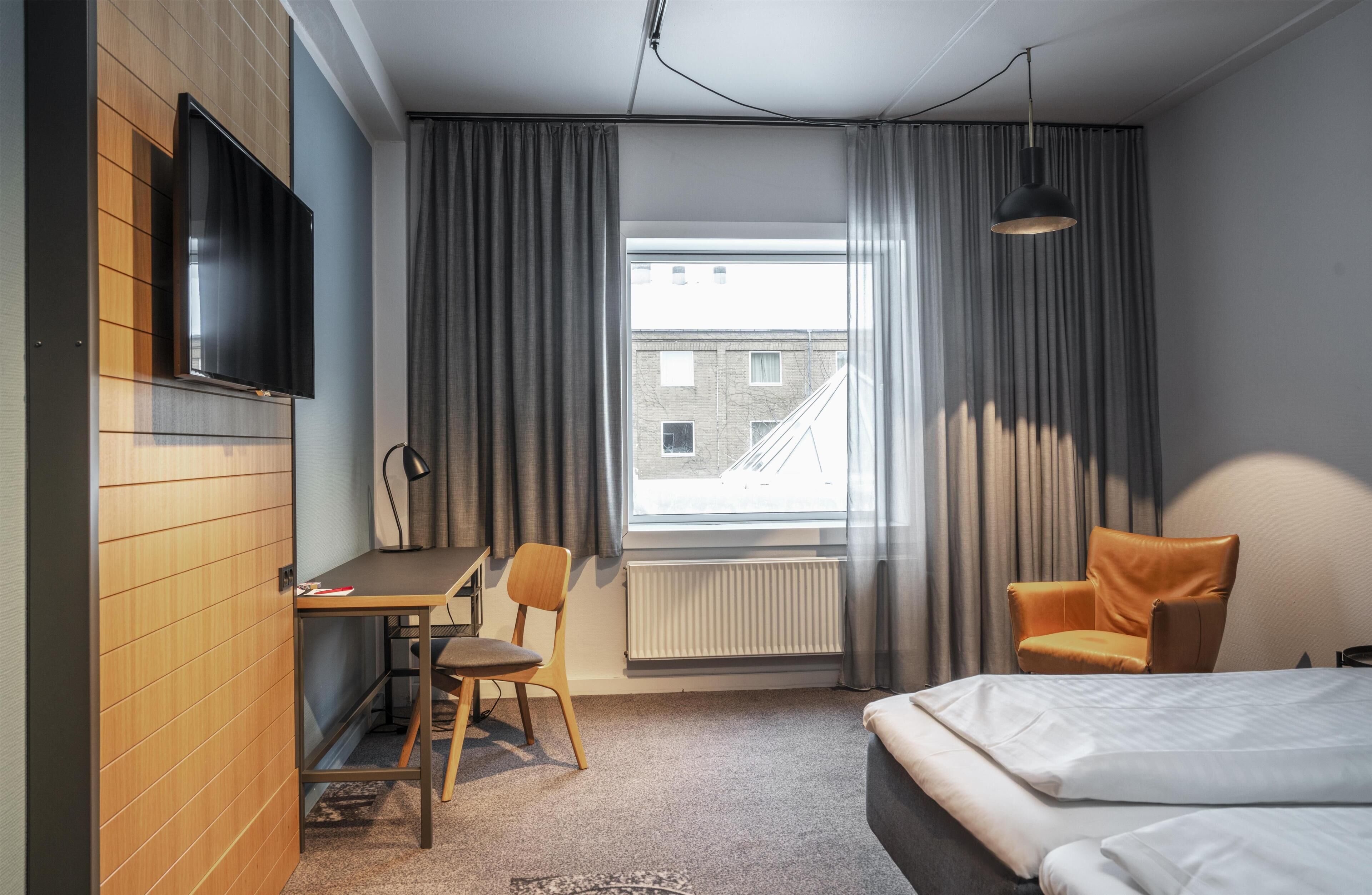 Une chambre d’hôtel avec un lit, un bureau, une chaise, une télévision et une fenêtre avec des rideaux.