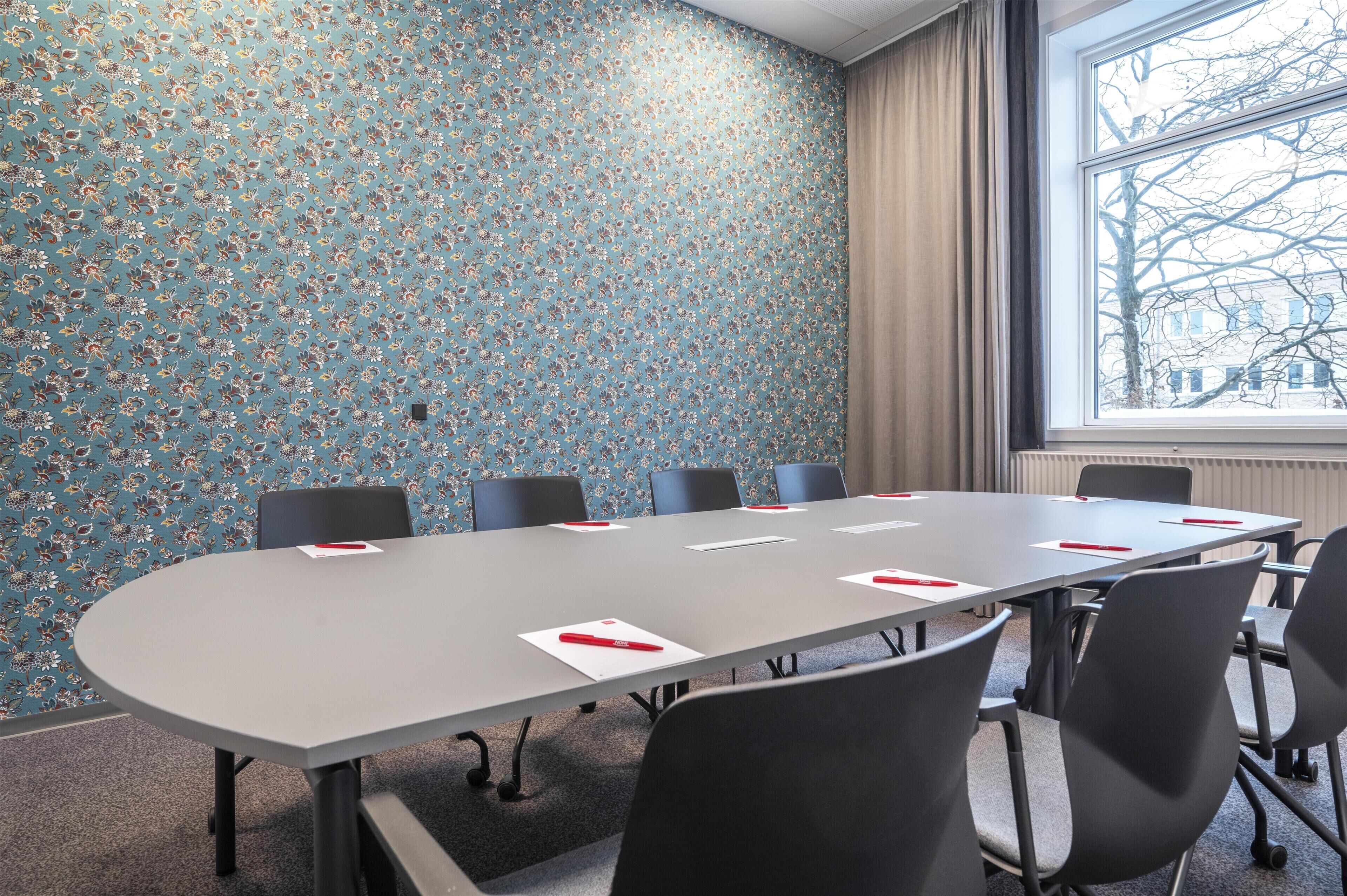 A conference room with a floral wallpaper, a large table with chairs, and a window with a view of trees and buildings.