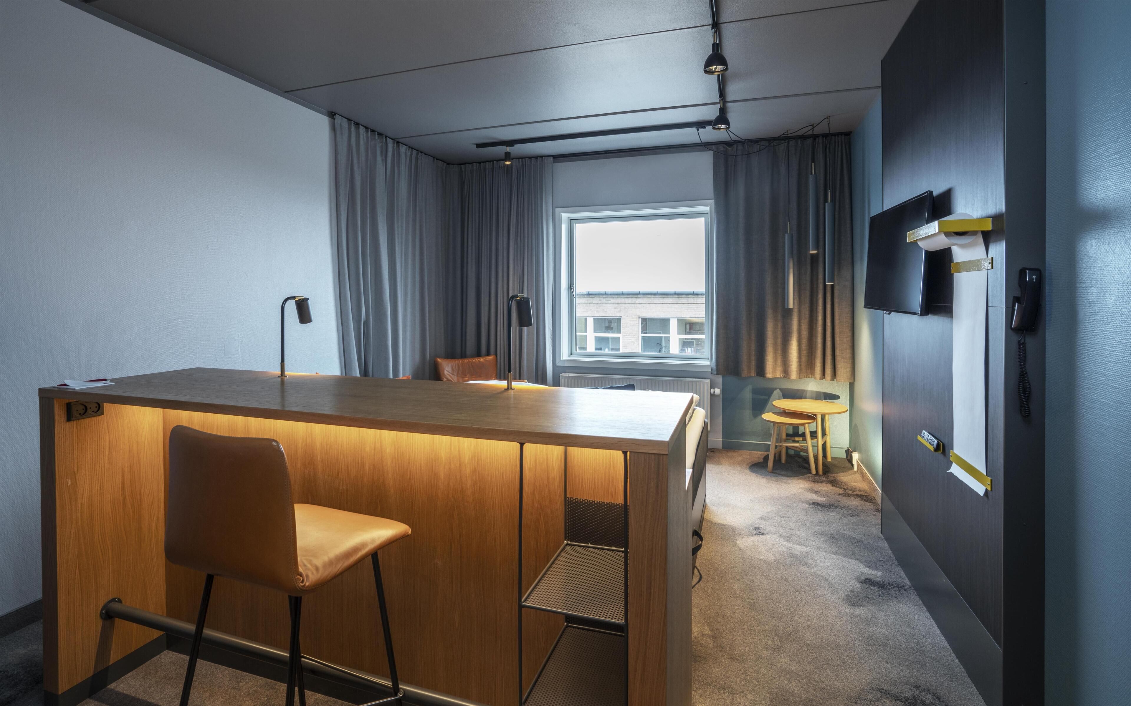 A hotel room with a wooden desk, two chairs, a small round table, a telephone, and a window with curtains.