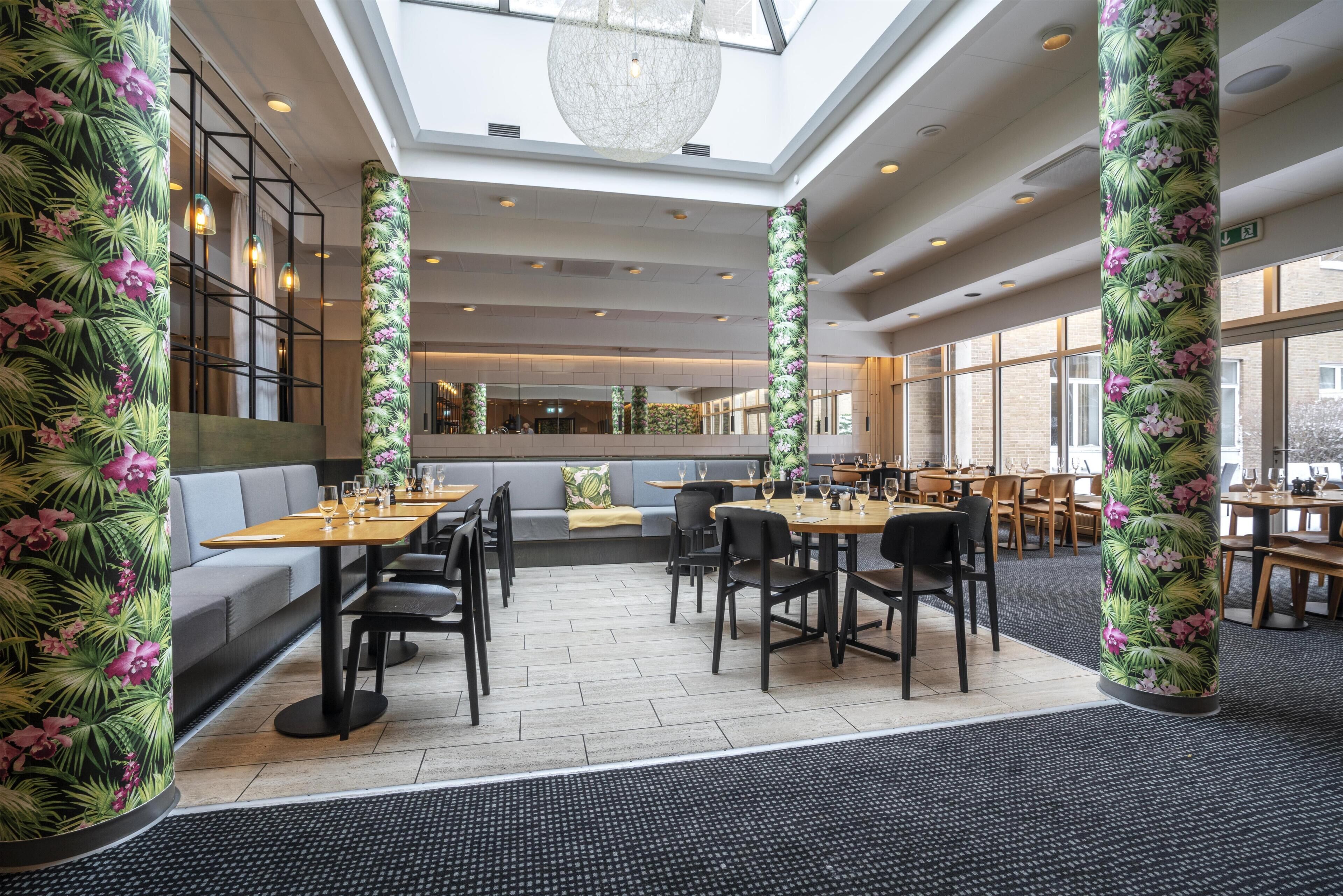 Restaurante com pé-direito alto, janelas amplas e colunas florais tropicais.