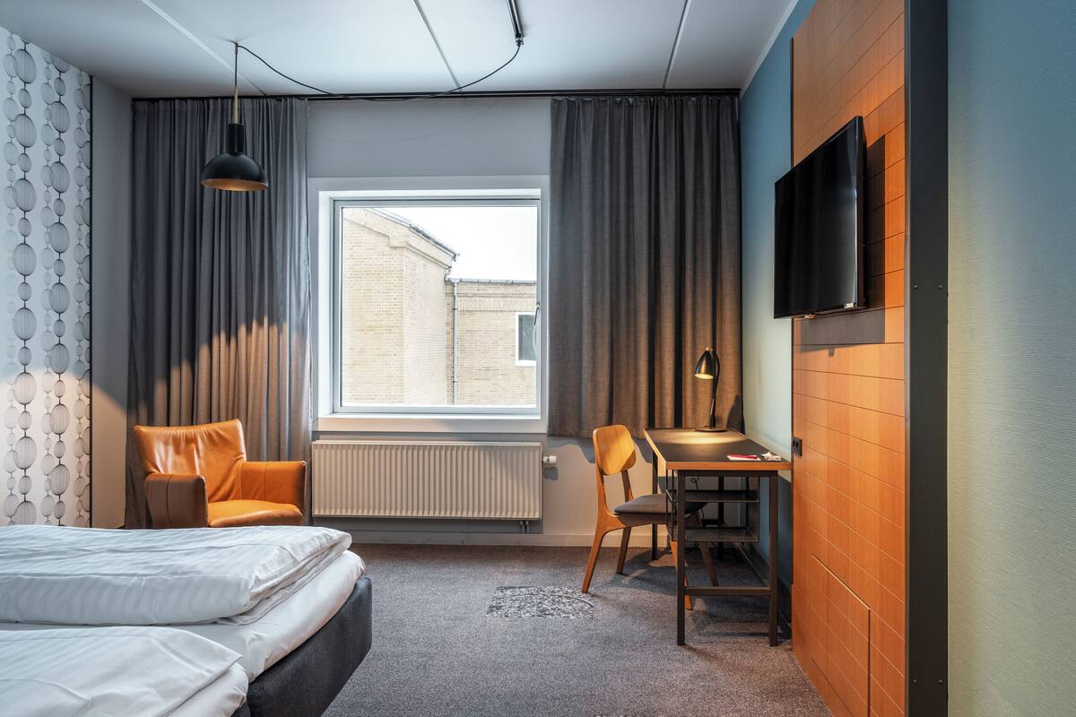 Et hotelværelse med en seng, to stole, et bord, et fjernsyn og et vindue med gardiner.